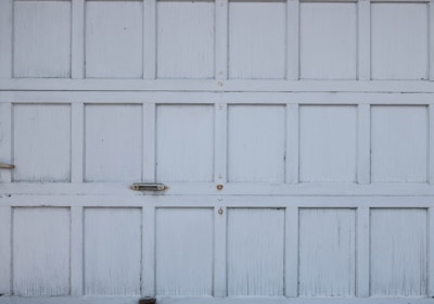 An old, weathered garage door with a pattern of rectangular panels. The panels have a white, slightly peeling paint finish. There are small metallic handles and fixtures visible on the surface.
