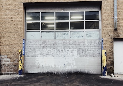 A large industrial garage door set into a tan brick building. The door is white with multiple small windows at the top, and there are yellow and blue protective bollards on either side. The ground in front of the door is paved and appears slightly worn.