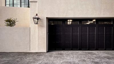 A modern garage door with a dark finish set against a light-colored stucco wall. A traditional-style lamp is mounted on the wall, and a small potted plant is visible on a ledge. The driveway is paved with patterned stone.
