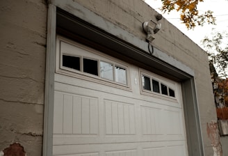 A large white garage door set in a beige concrete structure. Above the garage door, there are windows with three of the small panes being blacked out. A security light is mounted above the door. There are some visible signs of wear and paint coming off on the building and the surrounding autumn foliage is visible at the top right corner of the image.