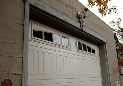 A large white garage door set in a beige concrete structure. Above the garage door, there are windows with three of the small panes being blacked out. A security light is mounted above the door. There are some visible signs of wear and paint coming off on the building and the surrounding autumn foliage is visible at the top right corner of the image.