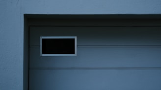 A garage door with horizontal panels and a small rectangular window located near the top left corner. Above the door, there's a section of textured brick wall illuminated by light, creating a contrast between the bright and shadowed areas.