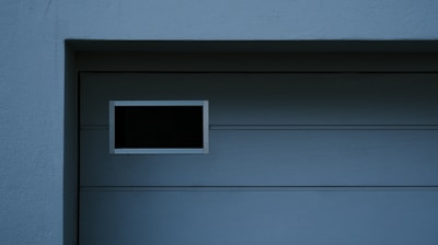 A garage door with horizontal panels and a small rectangular window located near the top left corner. Above the door, there's a section of textured brick wall illuminated by light, creating a contrast between the bright and shadowed areas.