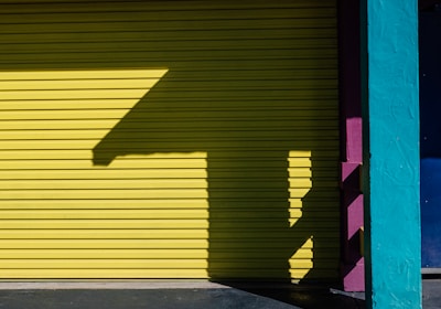 A brightly colored garage door with a bold yellow hue is flanked by a teal pillar and a section of purple under the red tiled roof. A distinctive shadow pattern is cast on the door, creating a geometric design.