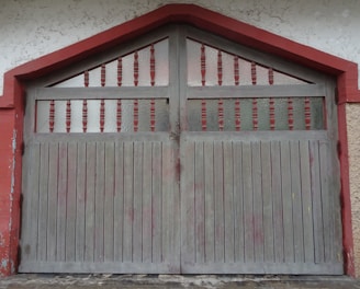A vintage-style garage door with vertical wooden slats and ornate red and frosted glass panels in the upper section. The frame is painted in red, contrasting with the gray of the door itself. The surrounding wall has a textured, stucco-like finish.