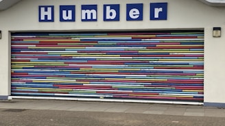 A decorative garage door with horizontal stripes in a variety of bright colors. Above the door is a sign with blue letters reading 'Humber'. The building facade is light-colored and the ground is paved.